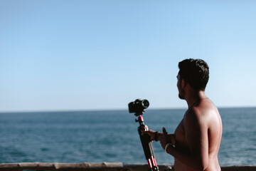 hombre joven sin camisa grabando video en la playa