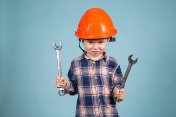 A cute funny little boy in an orange hard hat posing funny, holding wrenches