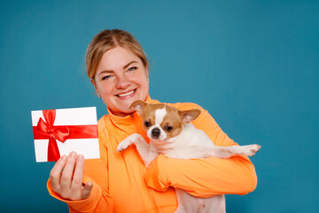 Happy young woman in orange t-shirt with Chihuahua dog holds gift certificate isolated on blue...