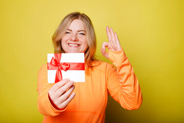 Fototapeta premium Happy young woman in orange t-shirt holds gift certificate isolated on yellow background. People lifestyle holidays concept.
