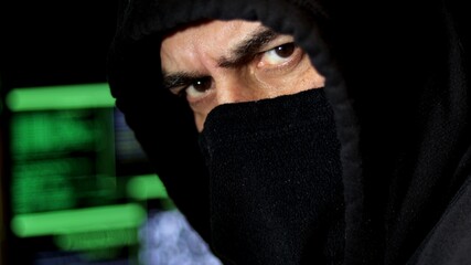 Close-up, cyber hacker with hood and covered face looks at the camera with serious look. Cyber terrorism concept.