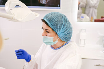 Side view photo of a woman dentist in uniform, medical mask and gloves ready begin to work with a patient in stomatology clinic.