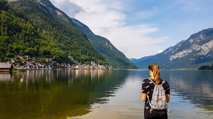 A girl with a small backpack standing at the edge of a lake in Hallstatt, Austria. Girl is looking at her phone. There is a small village on the other side of the lake. Serenity and calmness.