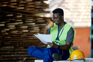 Smiling African American worker goes through reports during his coffee break at wood warehouse.