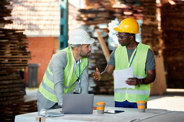 Black worker and warehouse inspector cooperate while going through distribution plans at storage...