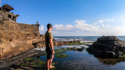 A man standing next to an edge of a cliffs, enjoying the waves splashing on cliffs, next to Tanah Lot Temple, Bali, Indonesia. Power of the nature. The man is enjoying his time and surrounding nature