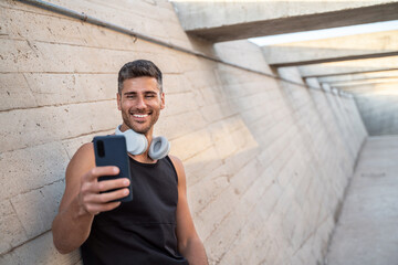 Attractive runner resting after training, standing on concrete wall background, using mobile phone, taking selfie.