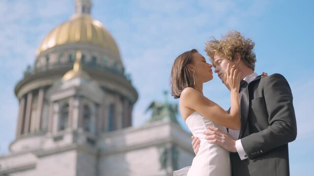Wedding photo shoot. Action. A couple poses next to St. Isaac 's Cathedral , a young man with long curly hair and his bride with bare shoulders and long earrings