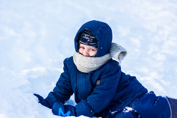 Fototapeta na wymiar A smiling happy child lies in a snowdrift on a sunny winter day. A lot of snow and very frosty. Active winter outdoor games.