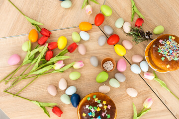 Fototapeta na wymiar Easter cake with sprinkles, painted eggs and flowers on wooden table, flat lay. Space for text