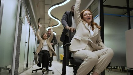 Excited young cheerful multiracial diverse colleagues having fun riding on office chairs in coworking space. Active happy millennial workers take a break enjoy play game laughing together at workplace
