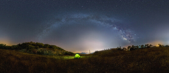Panorama of Milky Way bow with green illuminated tent at dry rocky landscape, Croatia