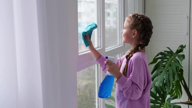 A little girl sprinkles water from a bottle on the window and wipes it off with a rag. Child dancing helps with cleaning the house. Clear the window.