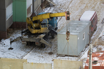 A crane manipulator installs a concrete fence at a construction site. The construction site is covered with snow. A manipulator crane is installed on a truck platform, next to a trailer for builders.
