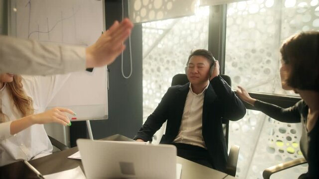 Young attractive businessman listening to music in headphones ignoring his colleagues. Confused office workers trying to interrupt him collegue for some tasks. High quality 4k footage