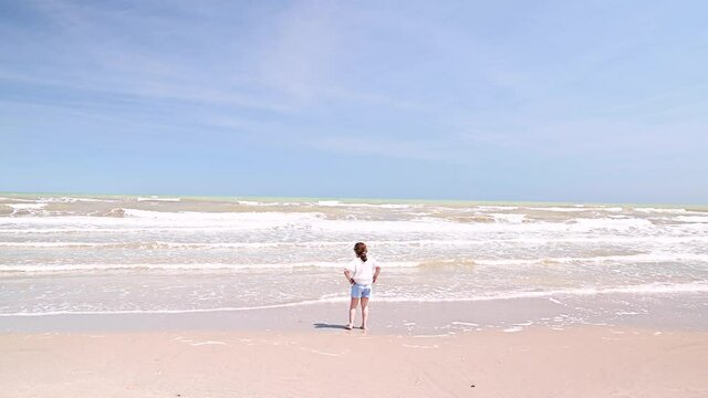 little girl walks along the beach in September. Blue sky ocean, waves and a child in summer clothes. Summer holiday concept in the south. High quality 4k footage