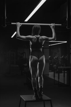A photo from behind of the sporty woman while she preparing to do wide-grip pull-ups in a gym. A muscular brunette girl wears a black top and high waist short shorts.