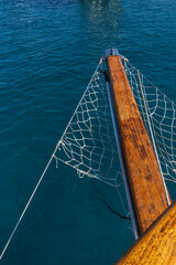 A wooden bowsprit of a sailboat with nets on the sides, over turquoise colored Mediterranean Sea.