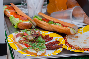 Sandwich with arugula, ham, cheese, jalapeno, sundried tomato and salami on plate on counter at summer local food market - close up. Gastronomy, street food, mediterranean and Italian dish concept