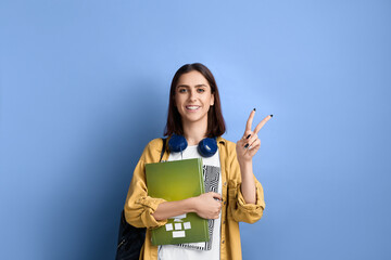 Good luck. Cheerful smiling student girl is holding books and making victory gesture with hand, confident with knowledge, wearing yellow shirt, white t-shirt, black bag and headphones over neck.