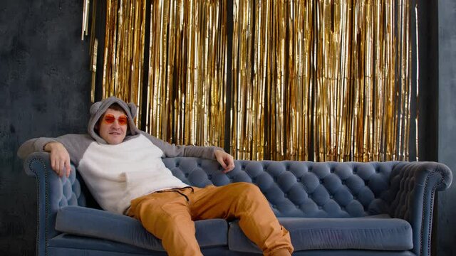 Serious and dissatisfied man in hood with ears and bright glasses sitting on sofa against wall with golden tinsel. Relaxed important male sprawled on couch and looking at camera.