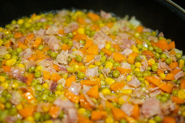 close up of a vegetable stir fry with carrots, corn, peas, onion and ham, fried in olive oil in a black frying pan, selective focus