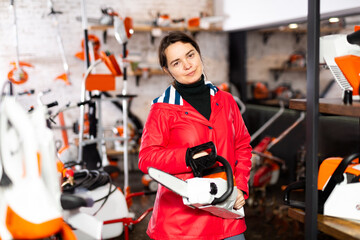 European woman standing in salesroom of gardening tools store with chainsaw in hands and looking in...