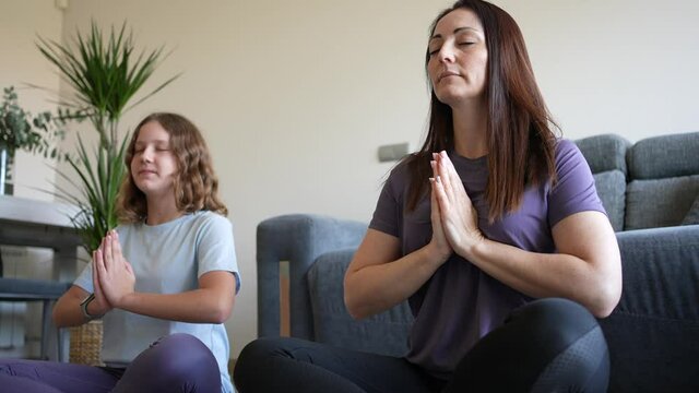 Preteen daughter meditating in lotus position with mother at home. Happy healthy family mother and teenage girl having fun doing yoga exercise relaxing together sitting on the floor at home.
