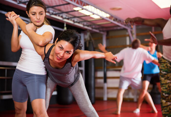 Latin and Asian women exercising armlock grip while sparring in gym. African-american man self-defence coach standing nearby and watching them.