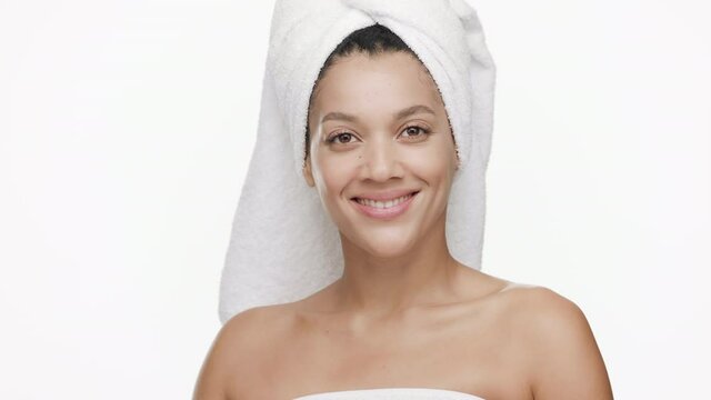 Slim pretty African American young woman with a towel on her hair and bare shoulders strokes her cheek and turns head to the camera smiling wide for it on white background | Skin moisturizing concept