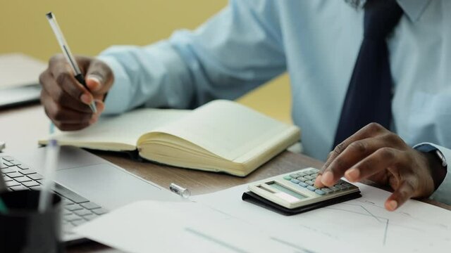 Male analyst or investor working on project and considering risks at table in modern office spbi. Close-up view of young businessman counts on calculator and writes in notebook, examines probabilities