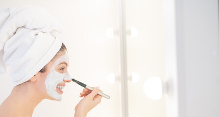 Cheerful woman with a towel on her hair and in a clay face mask fooling around with cucumbers in...