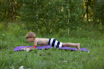 Thin boy in striped shorts does plank on fitness mat in clearing in forest. Child is engaged in physical exercises with fitness rubber bands in fresh air.