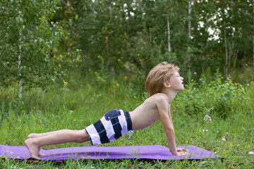 Little boy in striped shorts does plank exercise on fitness mat in park. Kid is engaged in physical exercises in nature.