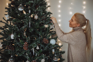 A young blonde girl with long hair in a beige dress decorates a Christmas tree at home