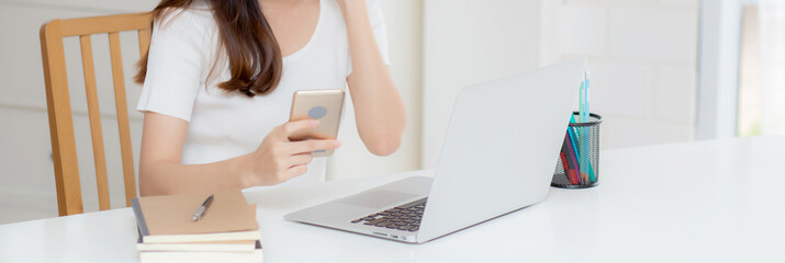 Young asian woman working laptop computer and reading smartphone on internet online on desk at home, freelance girl smiling using phone with social media, business and communication concept.