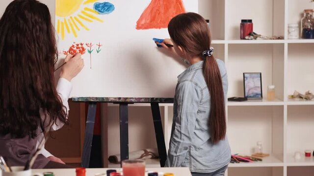 Art studio. Kids creativity. Painting lesson. Happy girl and woman drawing scenery picture together on white canvas in light room interior.