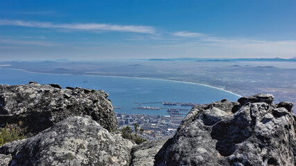 Fototapeta na wymiar There are huge grey boulders at the top of Table Mountain in Cape Town. In the gap between the stones, you can see the urban area, the gulf of the Atlantic Ocean. Blue sky. South Africa
