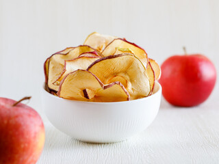 Dried thin slices, red apple fruit chips in white bowl and whole fruit on light background. Healthy...