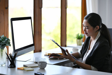 Businesswoman holding clipboard and working with computer in modern office.