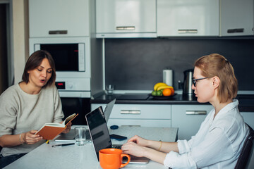 Woman's co-working, remotely work, home office, startup. Two attractive female freelancers sitting with laptops face to face in home kitchen.