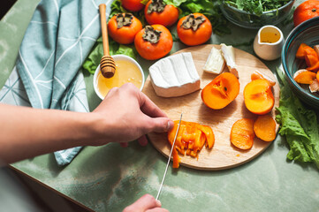 Ingredients and process of making a vitamin winter salad with persimmons, tangerines and cheese Female or teenage hand cuts persimmon on a round wooden board View from above Vegan lunch preparation.