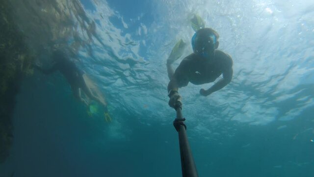 Young caucasian man swimming underwater freediving in mask and snorkel with fins taking pictures of himself on selfie stick and action camera