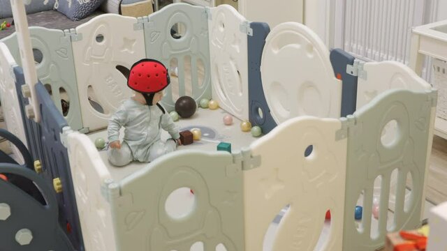 Cute toddler playing with soft montessori building blocks in baby playpen with play mat for babies and toddlers, 8 month old baby boy wearing safety helmet enjoy playing time in kids activity centre.
