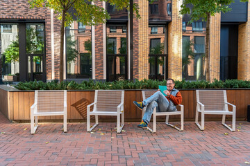 Millennial man with happy face checking phone sitting on one of four chairs in courtyard of modern  residential complex with landscaping. Portrait of excited man holding smart phone at street outdoors