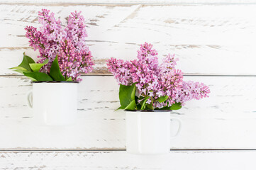 decorative composition for a country house. two white enameled mugs with branches of purple lilac on a white wooden table. flat look.