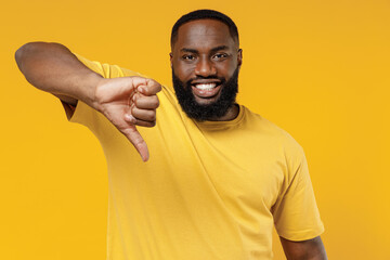 Young smiling dissatisfied displeased happy black man 20s in bright casual t-shirt show thumb down dislike gesture isolated on plain yellow color background studio portrait. People lifestyle concept.