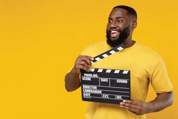 Young black man 20s wearing bright casual t-shirt holding classic black film making clapperboard look aside on copy space isolated on plain yellow background studio portrait. People lifestyle concept.