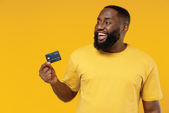 Young Smiling Satisfied Fun Rich Cool Happy Black Man 20s Wearing Bright Casual T-shirt Hold In Hand Credit Bank Card Look Aside On Workspace Isolated On Plain Yellow Color Background Studio Portrait.