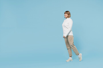 Full body side view elderly smiling cheerful fun caucasian woman 50s wear white knitted sweater walking going stroll isolated on plain blue color background studio portrait. People lifestyle concept.